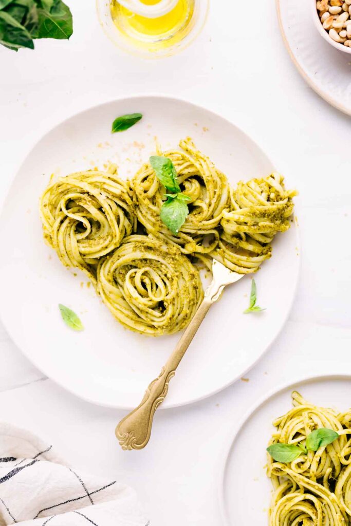 A light colored plate with three swirls of pasta al pesto on it and basil leaves as a garnish and a fork with pasta wrapped around it with another pasta plate in the right corner.