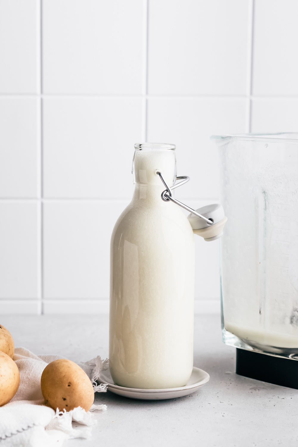 A milk bottle with potato milk next to a container from a blender with a few potatoes and a napkin and a white tile backdrop.