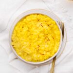 Yellow colored risotto in a shallow white bowl with little saffron threads as a garnish on a white napkin with a gold colored fork next to the bowl.