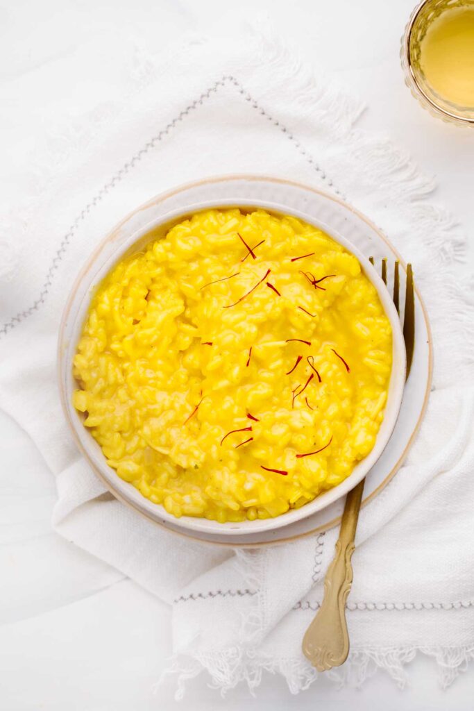 Yellow colored risotto in a shallow white bowl with little saffron threads as a garnish on a white napkin with a gold colored fork next to the bowl.
