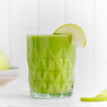 A green smoothie in a textured glass decorated with a slice of green apple on the rim of the glass on a white marble backdrop.
