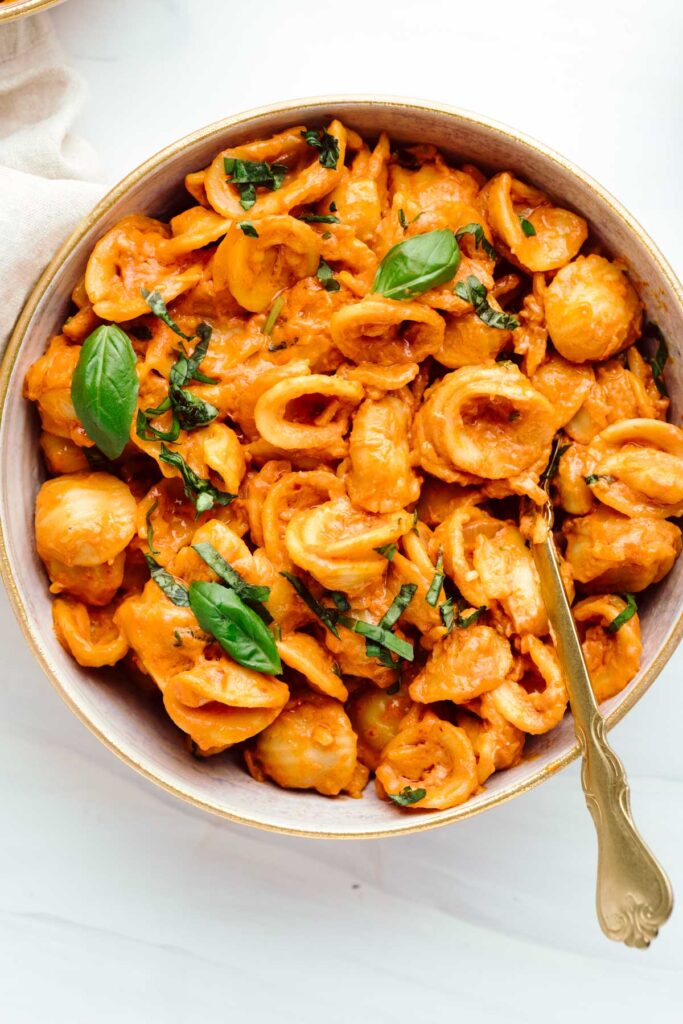 Orange colored shell pasta in a bowl with a gold fork in it and sprinkled with chopped basil and a few small basil leaves.