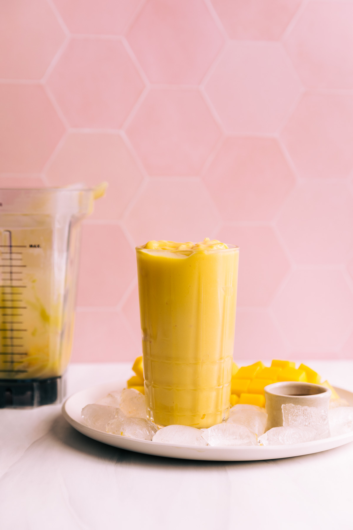 Mango lassi in a tall glass on a plate with ice cubes and fresh mango and a blender container with mango lassi in it in front of a pink tile backdrop