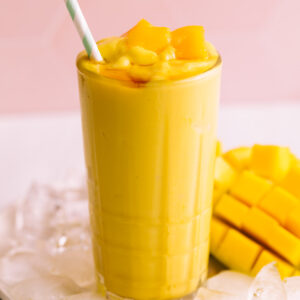 Mango lassi in a tall glass with a straw on a plate with ice cubes and fresh mango with a pink tile backdrop