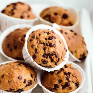 Chocolate chip muffins in white cupcake liners placed on each other on a white wooden tray