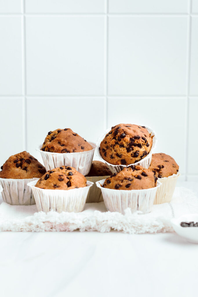 A white napkin with baked chocolate chip muffins placed on top of eachother in front of a white backdrop