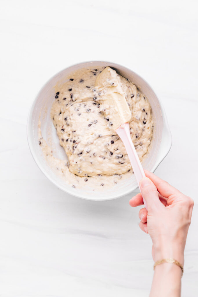 A white bowl with the ingredients for chocolate chip muffins in it with a hand with a spatula mixing it together