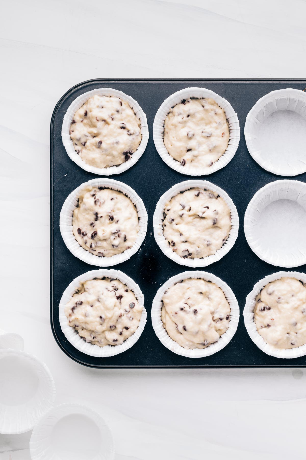 A black muffin tin pan with white cupcake liners and chocolate chip muffins batter in it