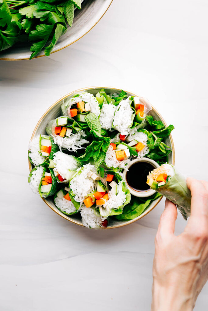A bowl with summer rolls sliced diagonally and presented to show the inside on a white backdrop with a hand dipping a roll in soy sauce