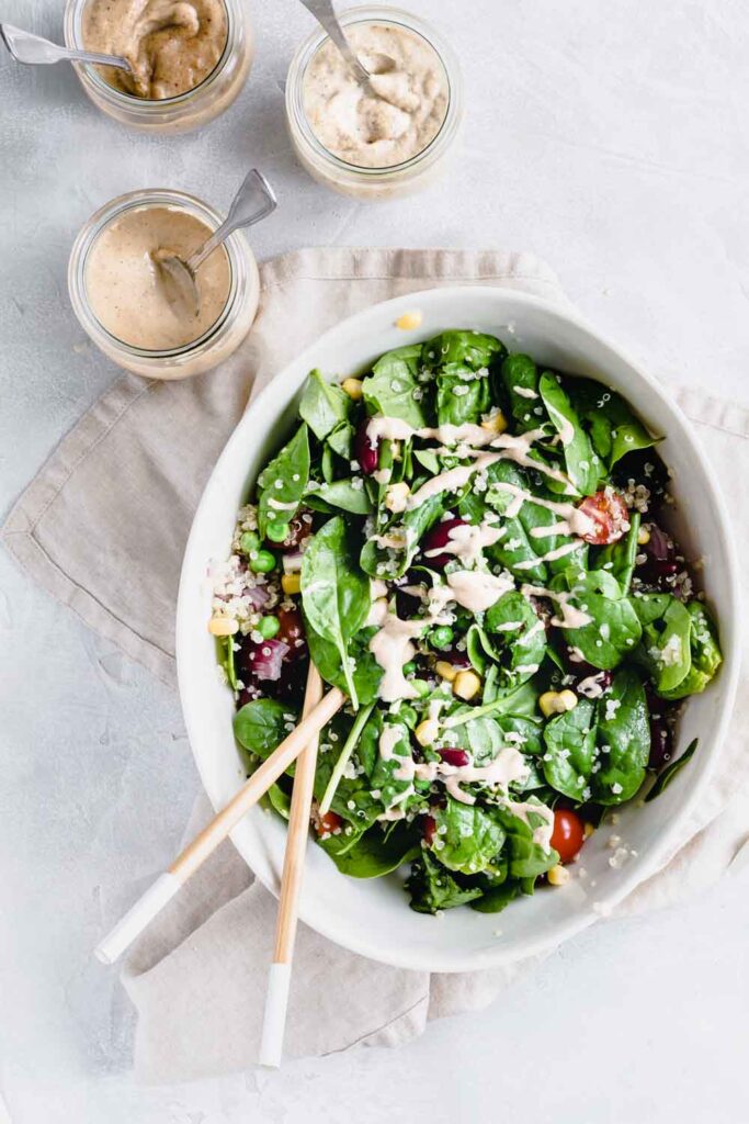 Quinoa salad in oval bowl with chopstick on a lightbrown napkin and a light backdrop next to a small jar of dressing with a teaspoon.