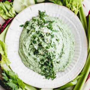 white bean dip in bowl with green herbs as topping on a backdrop of raw vegetables