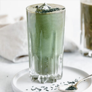green smoothie in tall glass on white plate with a spoon with spirulina