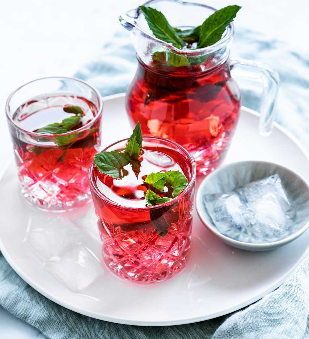 two glasses red cold hibiscus tea with fresh mint leaves next to a small bowl with ice cubes and a small carafe with hibiscus tea and mint leaves on white plate