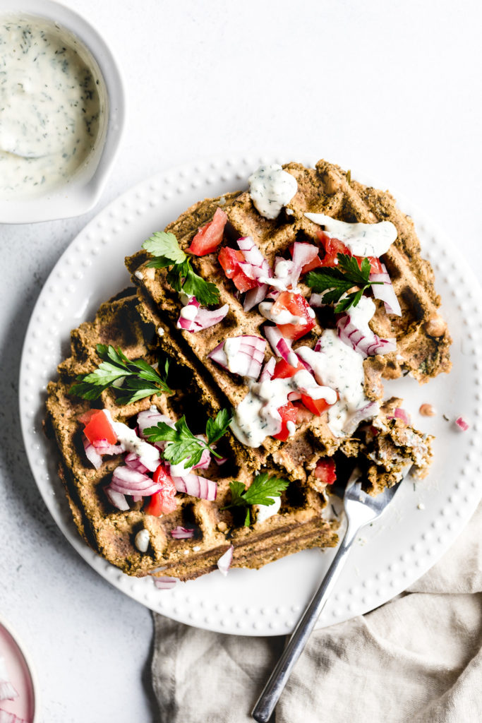 Falafel waffle on a white plate with a fork topped with fresh herbs, onion and sauce