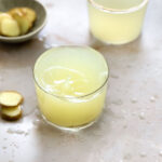 yellow ginger shot drink with splash in a small glass on a light brown backdrop with a glass with ginger juice in the background and fresh ginger