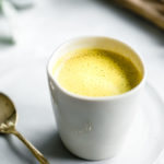 golden milk in white cup on a small white plate next to a golden teaspoon