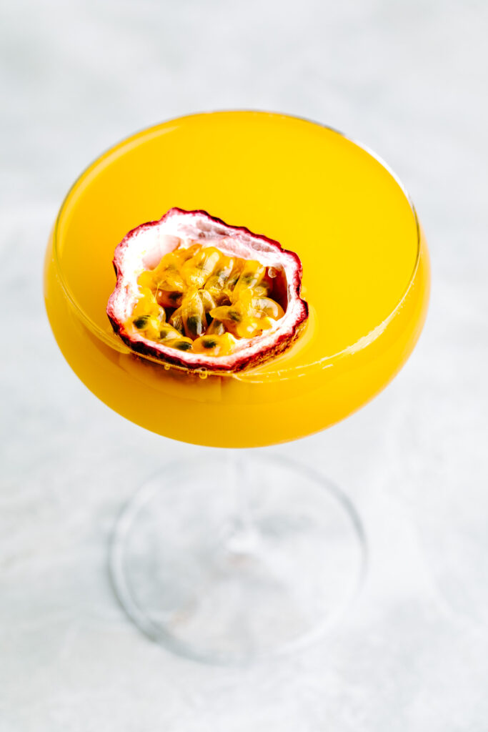 Orange colored drink Pornstar Martini Mocktail in a Martini glass with half a passion fruit floating on top on a light grey backdrop